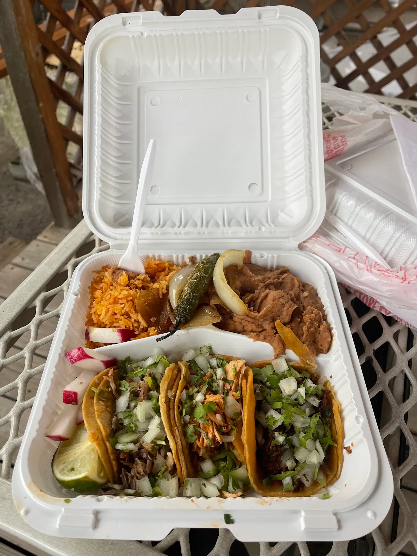 A takeout plate with three tacos, Mexican rice, refried beans, a grilled jalapeno, radish, and lime